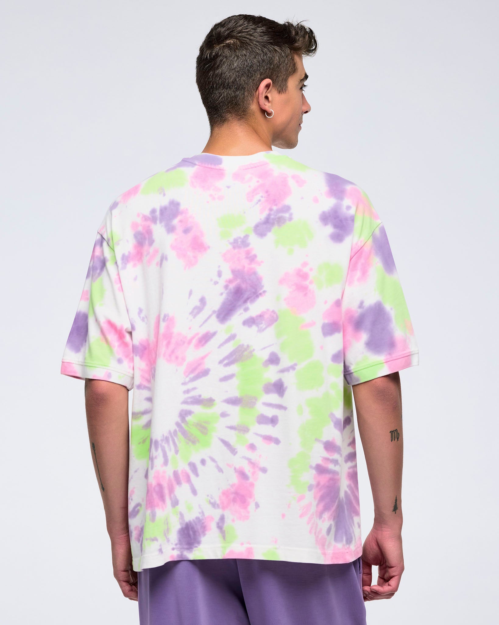 Back view of man standing against a white background, looking to the side, wearing an oversized pastel tie-dye pattern T-shirt with a large purple “ZUMBA” logo and purple pants.