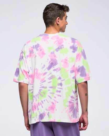 Back view of man standing against a white background, looking to the side, wearing an oversized pastel tie-dye pattern T-shirt with a large purple “ZUMBA” logo and purple pants.