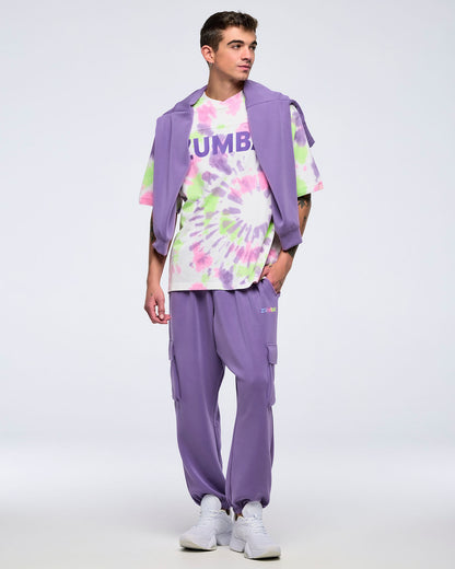 Full-body view of a man on a white background wearing a pastel patterned tie-dye “ZUMBA” T-shirt, a purple sweatshirt draped over his shoulders, purple cargo jogger pants, and white sneakers.