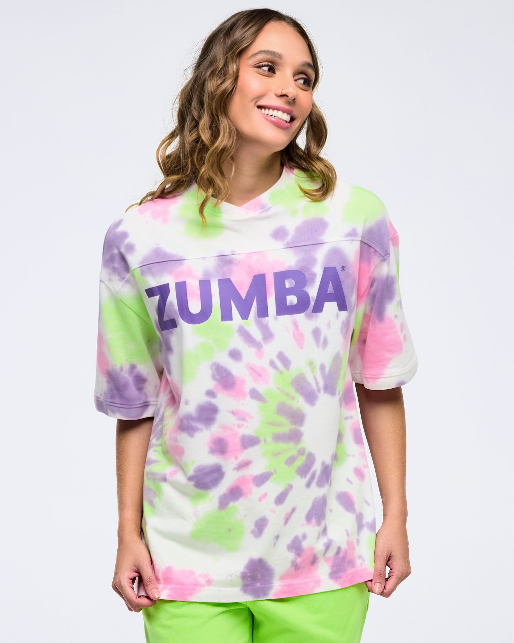 Smiling woman on a white background wearing an oversized pastel patterned tie-dye “ZUMBA” T-shirt and bright lime green pants, looking off to the side.