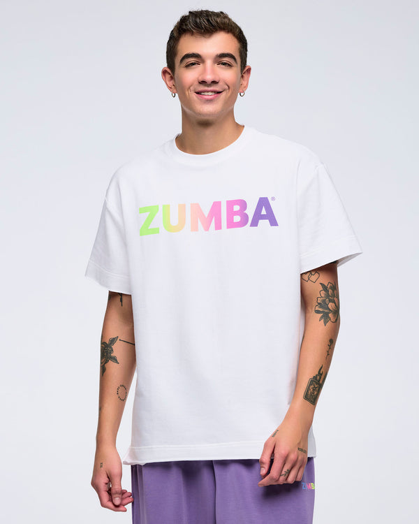 Smiling man on a white background wearing a white T-shirt with a pastel rainbow “ZUMBA” logo and purple pants.