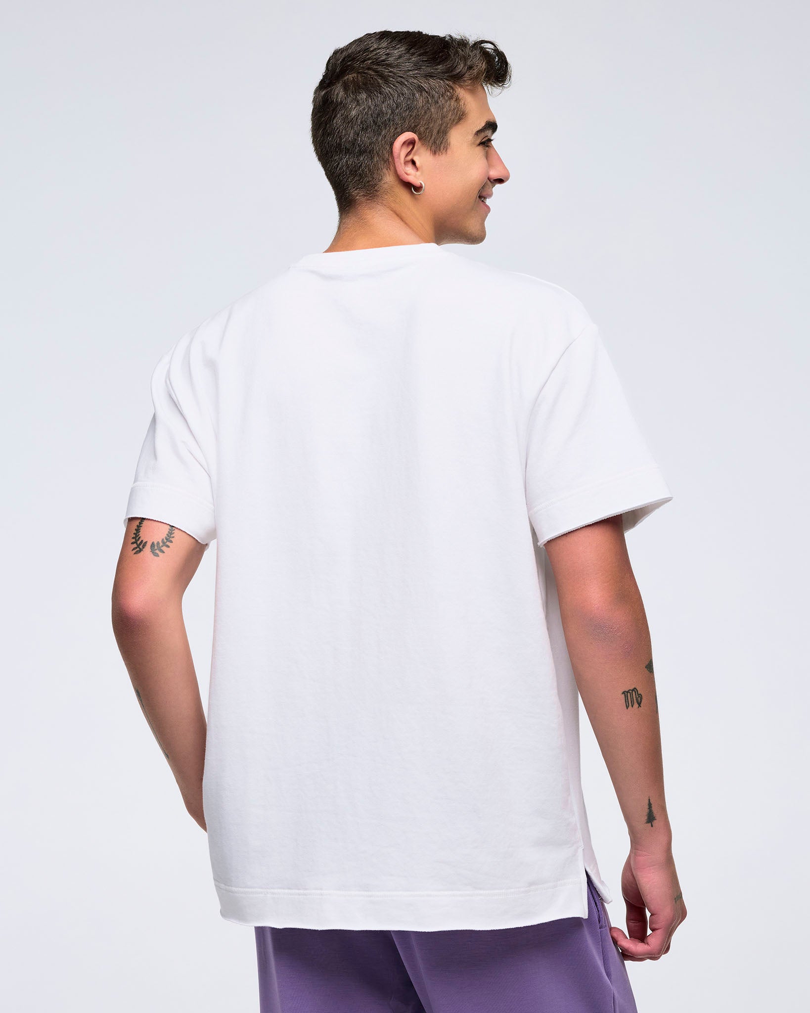 Back view of a man on a white background wearing a plain white T-shirt and purple pants, turned slightly to the side.