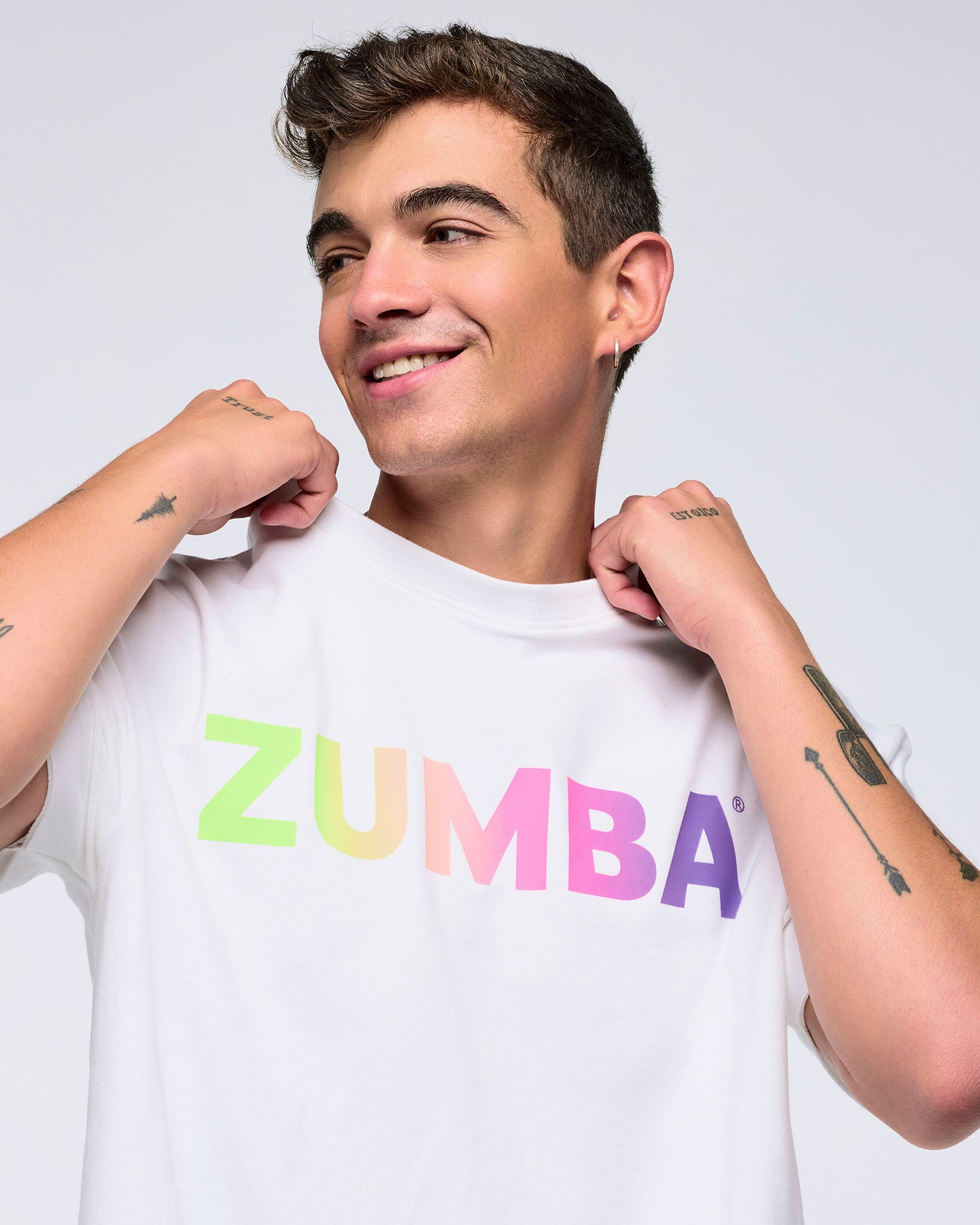 Close-up of a smiling man on a white background adjusting the collar of a white T-shirt with a pastel rainbow “ZUMBA” logo.
