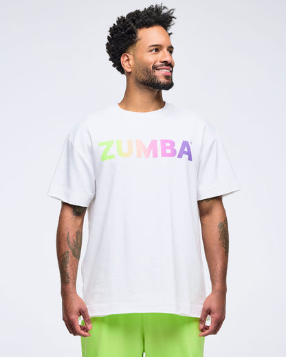 Smiling man on a white background wearing a white T-shirt with a pastel rainbow “ZUMBA” logo and bright lime green pants, looking off to the side.