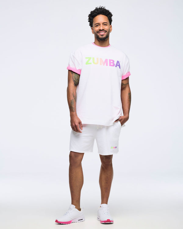 Full-body view of a smiling man on a white background wearing a white “ZUMBA” T-shirt layered over a pink shirt, white shorts, and white sneakers with pink accents.