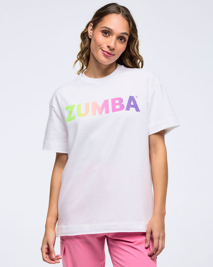 Smiling woman on a white background wearing a white T-shirt with a pastel rainbow “ZUMBA” logo and pink pants.