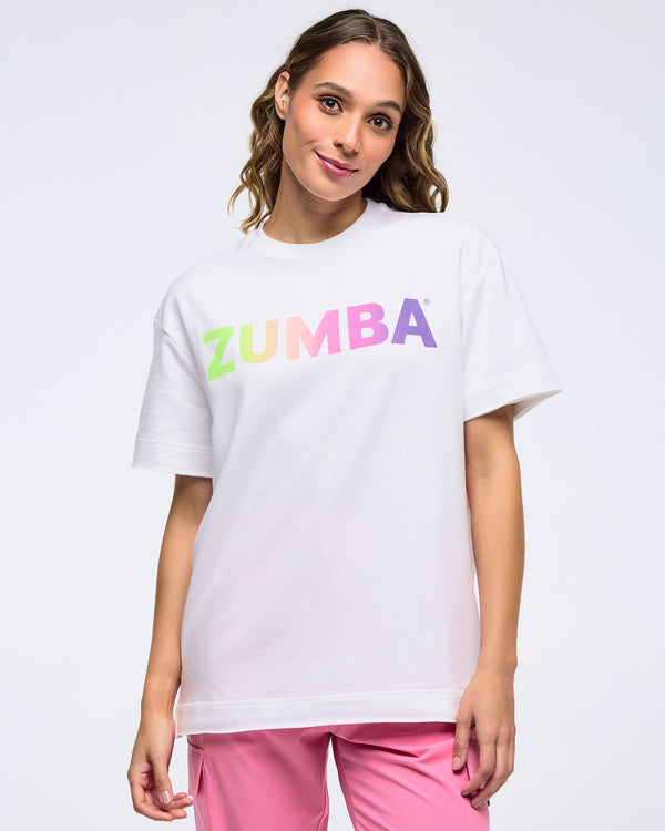 Smiling woman on a white background wearing a white T-shirt with a pastel rainbow “ZUMBA” logo and pink pants.