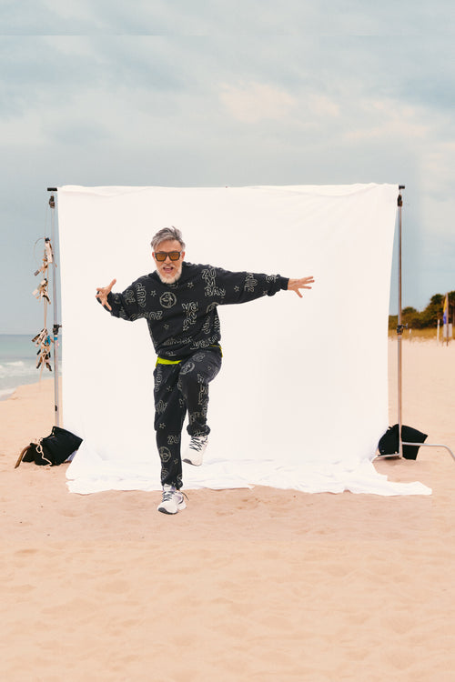 Beto (founder of Zumba), poses in front of a white backdrop wearing a black graphic sweatshirt and matching black joggers, white shoes and in sunglasses.