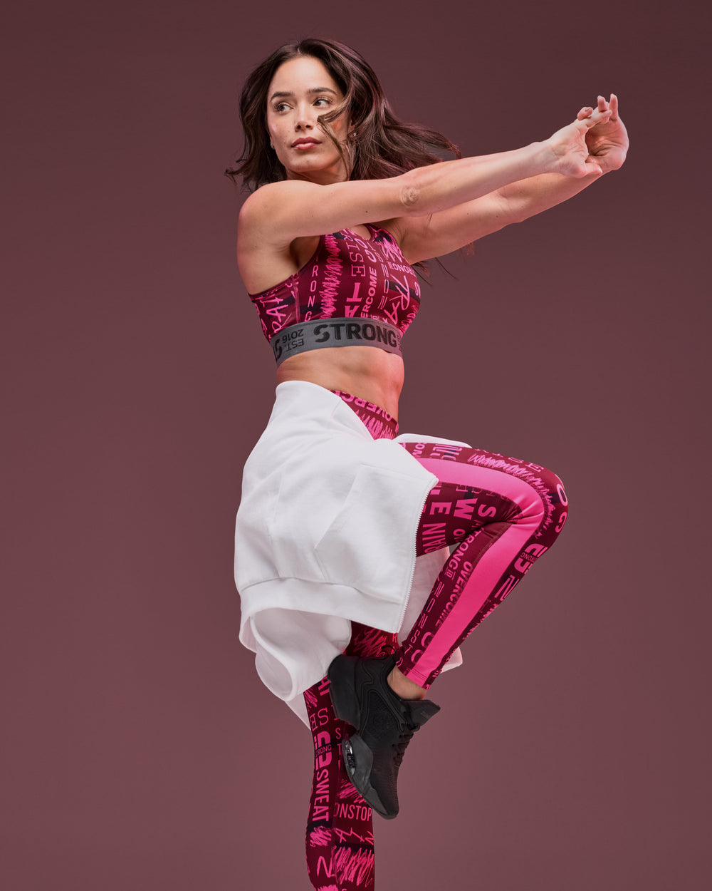 Banner featuring a female model in a pink graphic activewear set against a mauve studio background. She wears a matching sports bra and high-waisted leggings with bold text and abstract lettering in pink and burgundy, paired with black athletic shoes. A white hoodie is tied around her waist, and the gray elastic bra band reads “STRONG.” The model balances on one leg with arms extended, highlighting the coordinated workout outfit in motion.