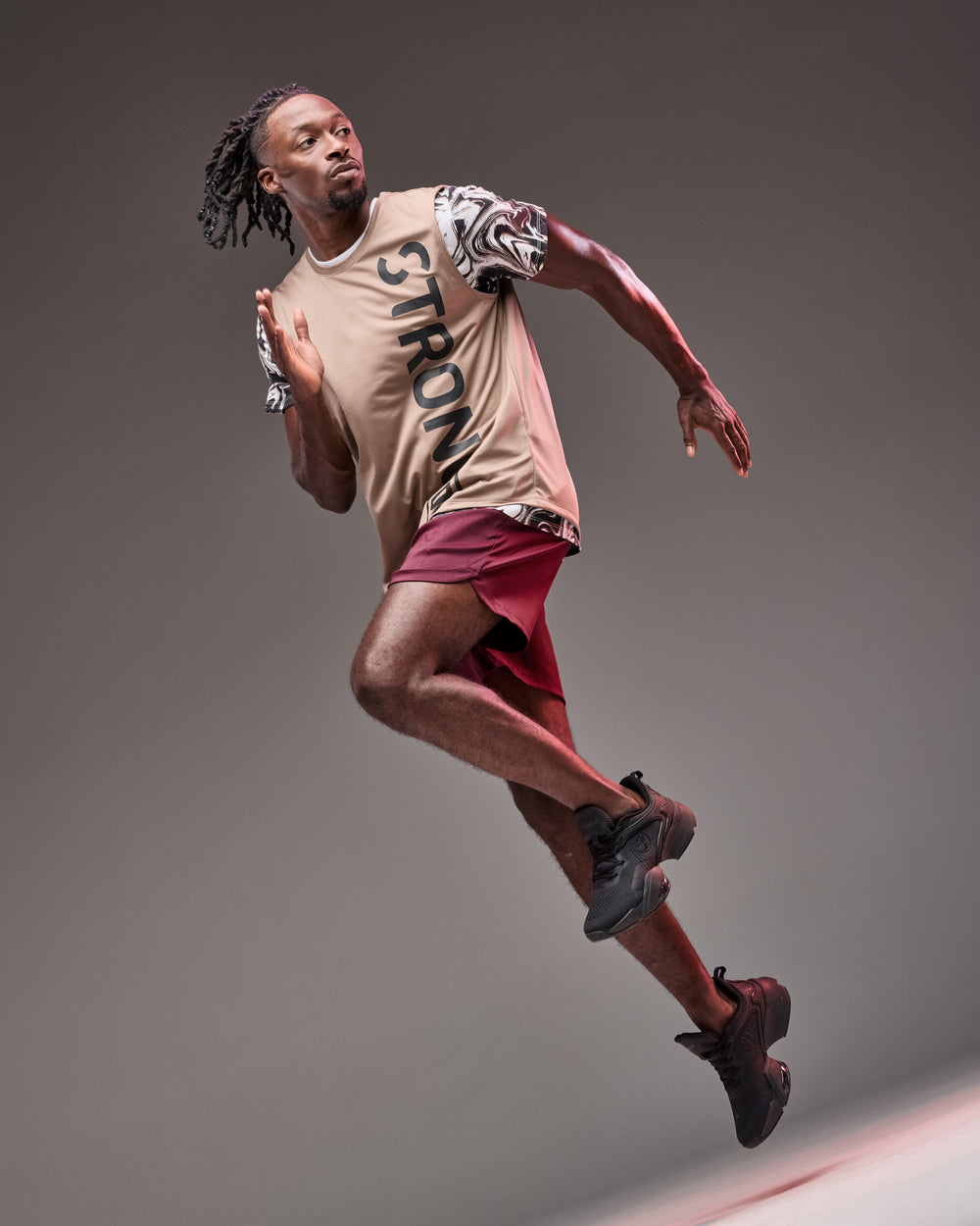 Banner featuring a male model in motion against a gray studio background. He wears a beige short-sleeve athletic T-shirt with marbled black-and-white sleeves and hem, plus bold vertical “STRONG” lettering down the front, paired with burgundy training shorts and black athletic shoes. The dynamic jumping pose highlights the lightweight, sporty outfit.