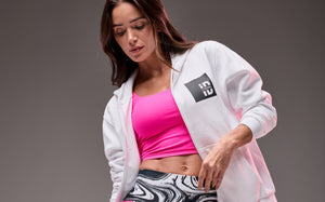 Banner featuring a female model in layered activewear against a gray studio background. She wears a white zip-up hoodie with a black “STRONG iD” logo patch over a bright pink fitted crop top, paired with high-waisted leggings in a black, white, and pink marbled print. The jacket is worn open to highlight the sporty, coordinated set.