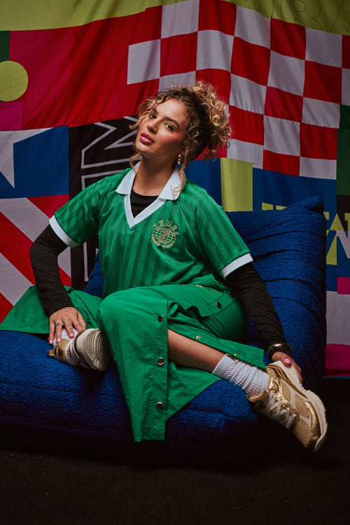 A female model in a green striped Zumba polo and gold sneakers lounges on a blue chair before vibrant flag backdrops.