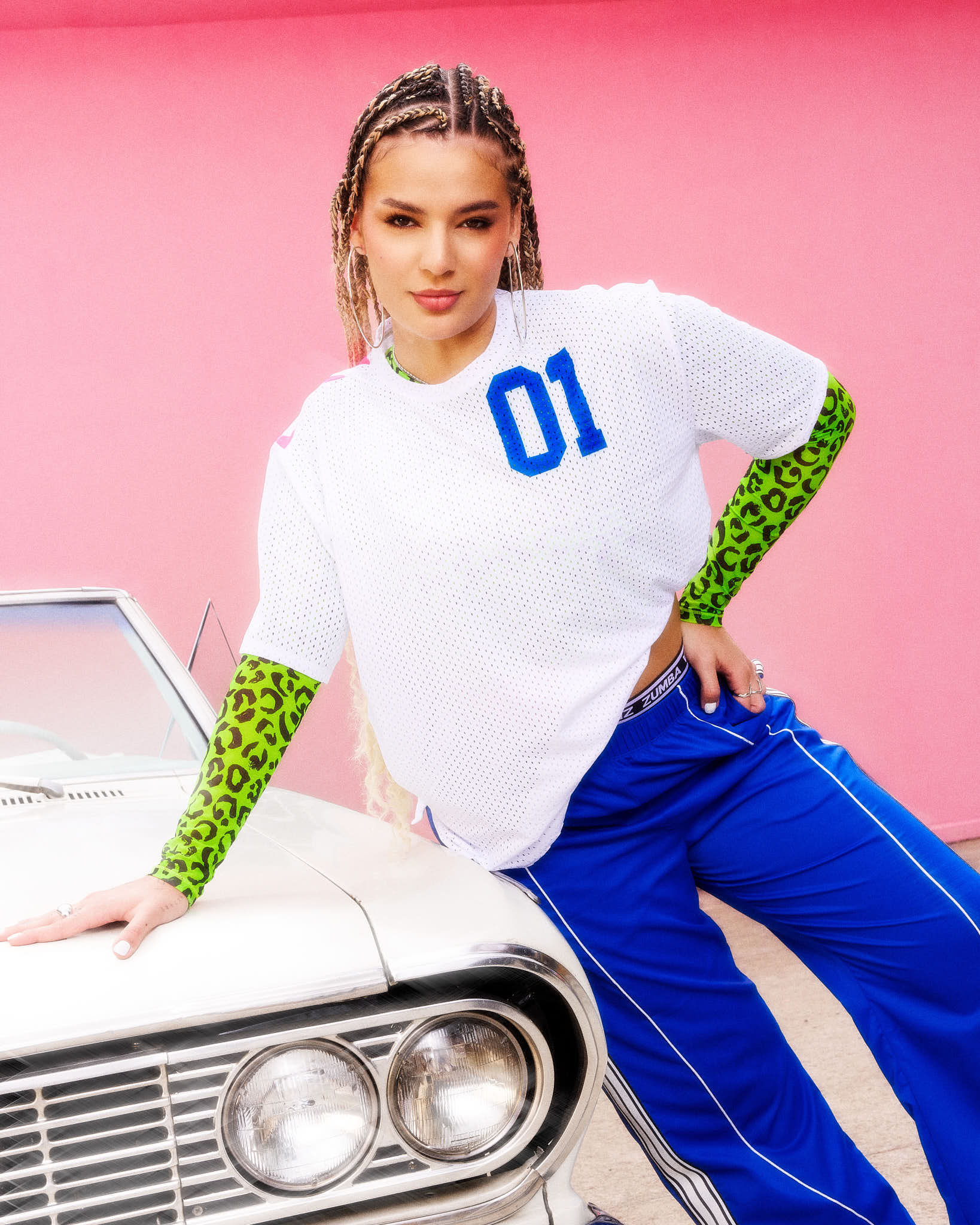A female model posing in a white mesh short-sleeve jersey top with blue '01' lettering, paired with bright blue track pants over a green leopard-print long sleeve top, leaning against a white classic car in front of a pink background.
