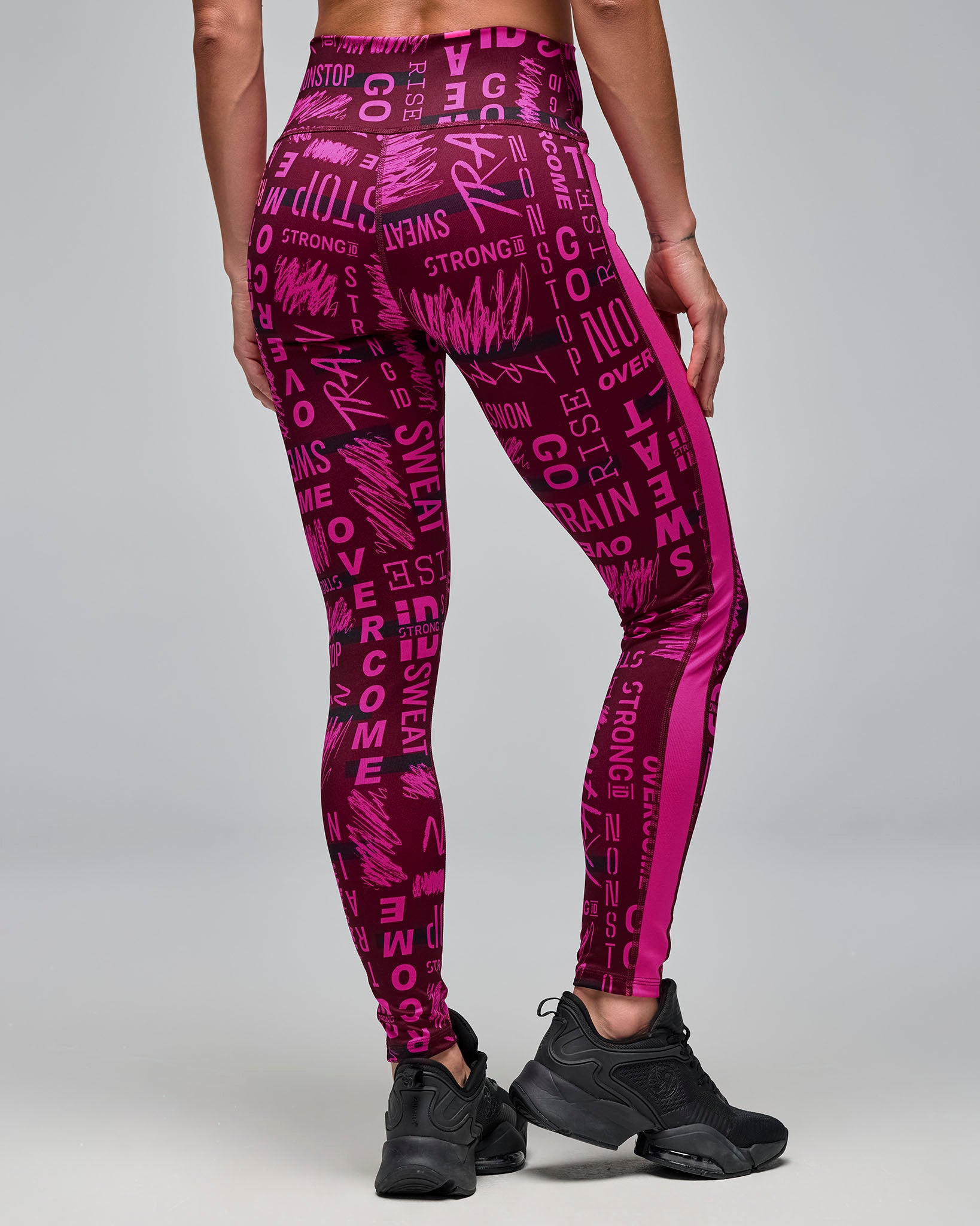 Back view of burgundy and pink high-waisted ankle leggings with an allover motivational word print and bright pink side panels. Female model wears the fitted leggings with black athletic sneakers against a light gray background.