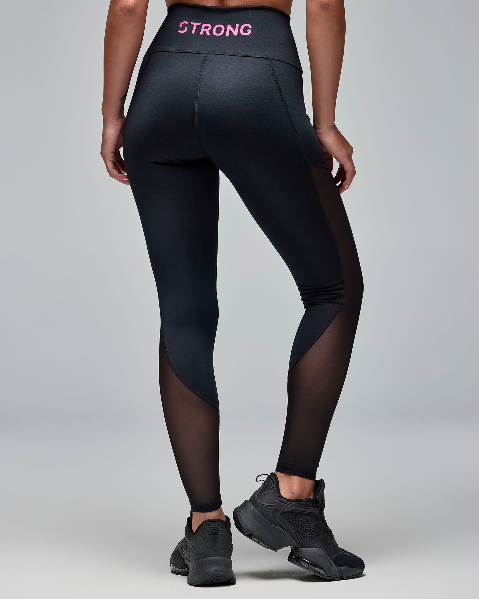 Back view of black high-waisted leggings with sheer mesh side panels and pink “STRONG” text on the waistband. Female model wears the full-length leggings with black athletic sneakers against a light gray background.