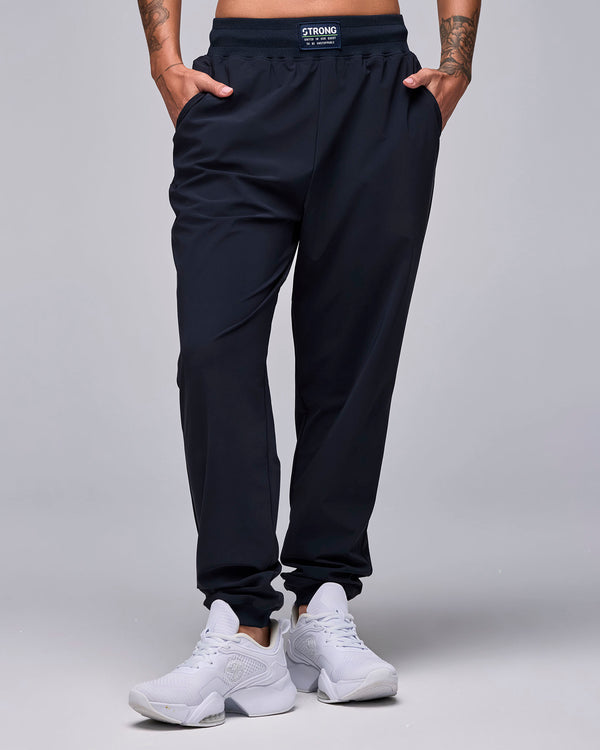 Female model in the black high-waisted joggers, with a closer look at the waistband label: white 'STRONG' text over the tagline 'UNITED IN OUR QUEST TO BE UNSTOPPABLE.'