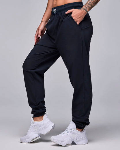 Female model in black high-waisted joggers with a relaxed fit, slanted side pockets, elastic cuffs, and a black waistband label with white 'STRONG' text, shown in side view with one hand in the pocket and paired with white shoes.