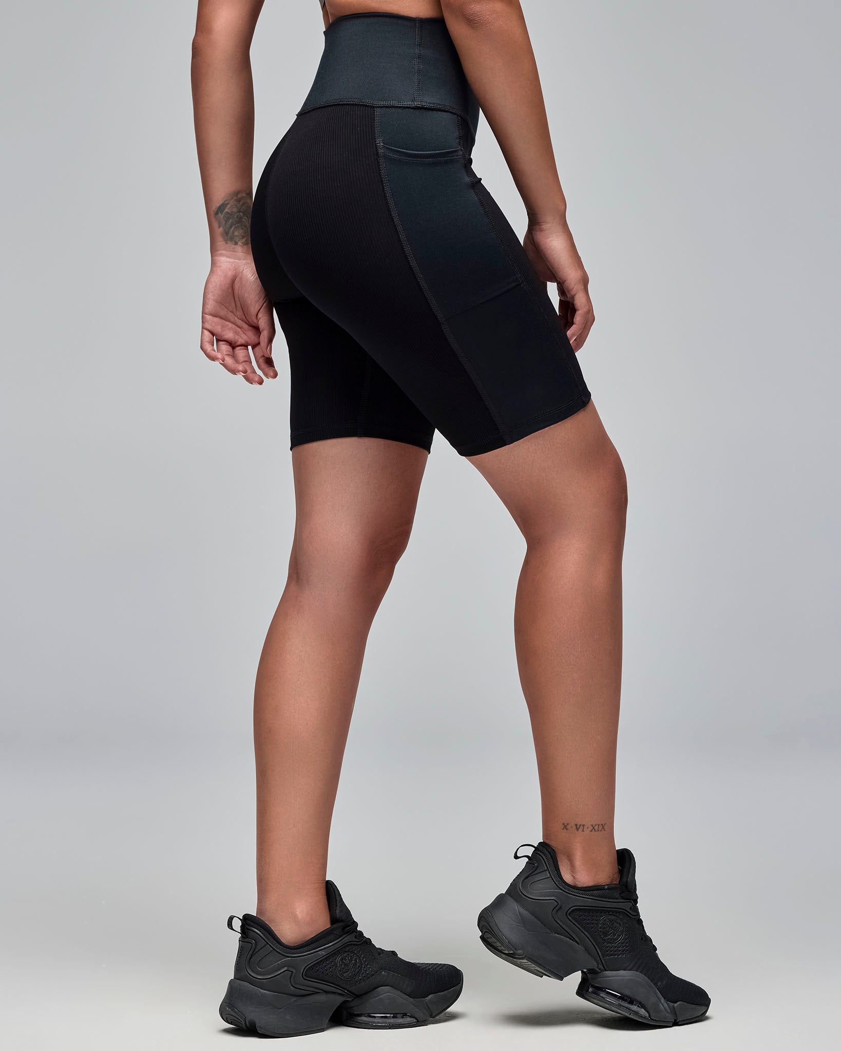 Side and back view of black high-waisted biker shorts with ribbed texture panels and side pockets. Female model wears the fitted mid-thigh shorts with black athletic sneakers against a light gray background.
