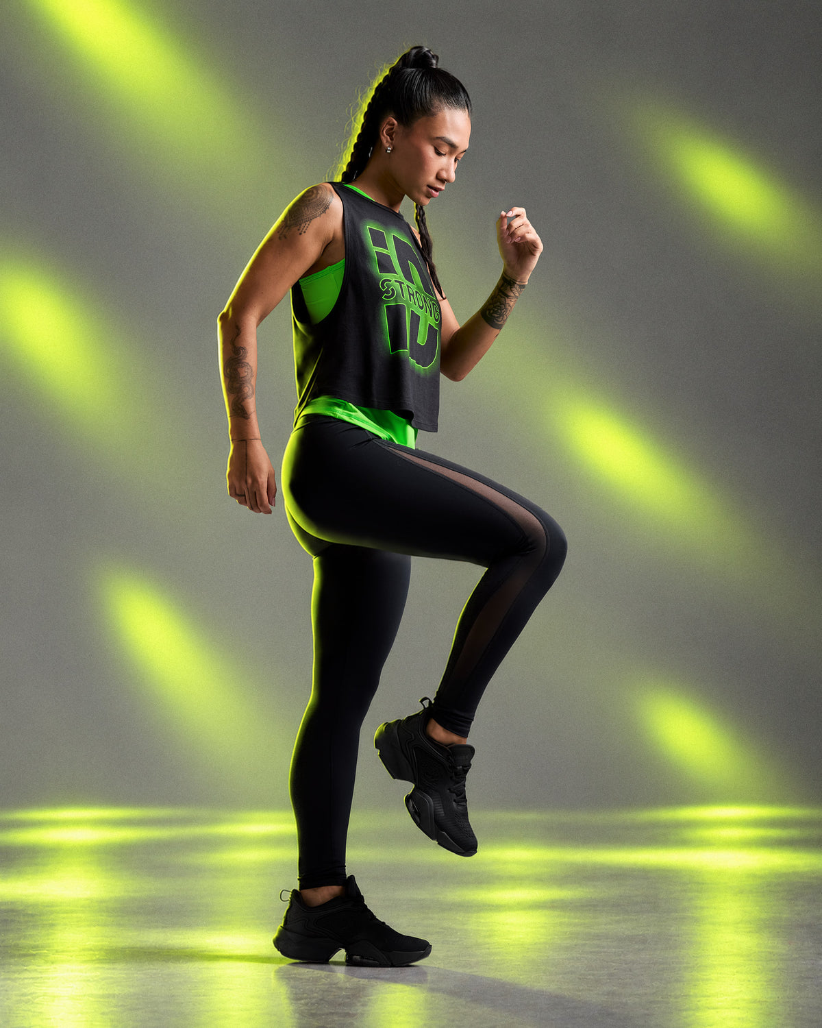 Female model in the black mesh insert leggings, mid-step with one knee raised, wearing a black 'STRONG iD' crop muscle tank over a neon green top. Neon light beams on a grey backdrop.