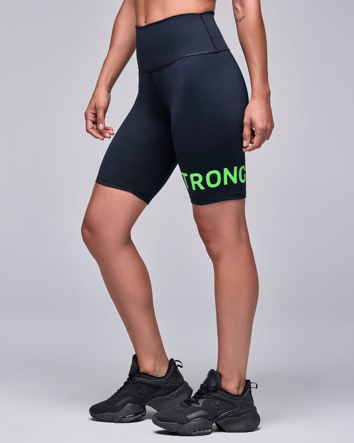 Black high-waisted biker shorts on a female model, shown in side view, with the neon green 'STRONG' text wrapping the left leg.
