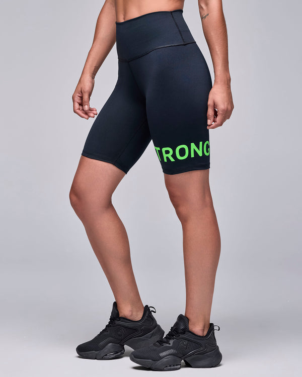 Black high-waisted biker shorts on a female model, shown in side view, with the neon green 'STRONG' text wrapping the left leg.