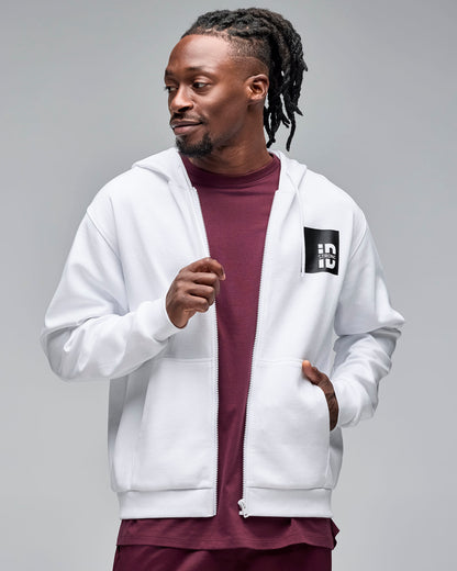 White zip-up hoodie with drawstrings and a black chest logo patch. Male model wears the long-sleeve hoodie open over a burgundy crewneck top against a light gray background.