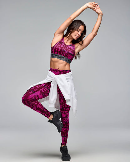 Burgundy and pink graphic-print sports bra with a gray elastic under-band, shown on a female model wearing matching high-waisted leggings, a white hoodie tied around the waist, and black sneakers in a dynamic pose.
