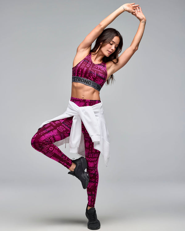 Burgundy and pink graphic-print sports bra with a gray elastic under-band, shown on a female model wearing matching high-waisted leggings, a white hoodie tied around the waist, and black sneakers in a dynamic pose.