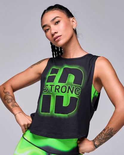 Female model in a black crop muscle tank with a large graphic: oversized black 'iD' letters with a glowing neon green outline and 'STRONG' across the middle. Paired with neon green and black swirl print leggings.