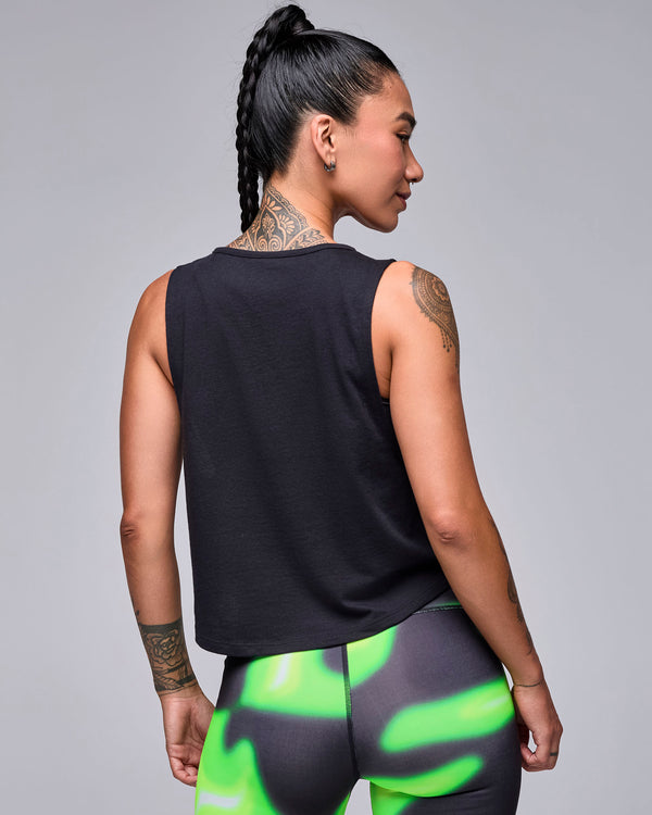 Shown from the back, the black crop muscle tank, plain on the back with a rounded hem, over neon green and black swirl print leggings.