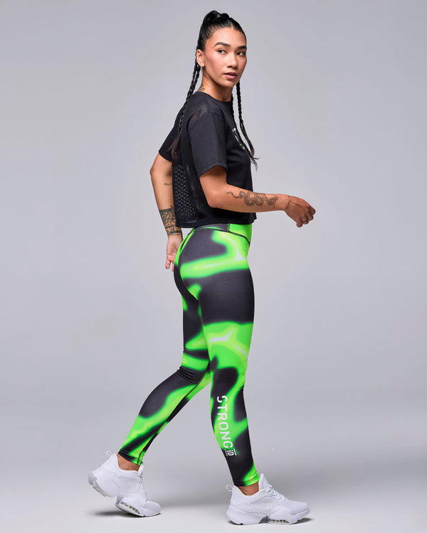 Female model walking in the black 'STRONG' boxy top, shown in side view with the mesh back visible, plus neon green and black swirl print leggings and white shoes.