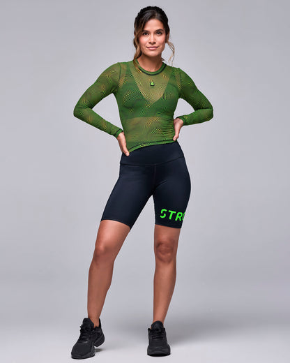 Full view of a female model in the wavy check print mesh long sleeve top with black 'STRONG iD' biker shorts and black shoes, hands on hips.