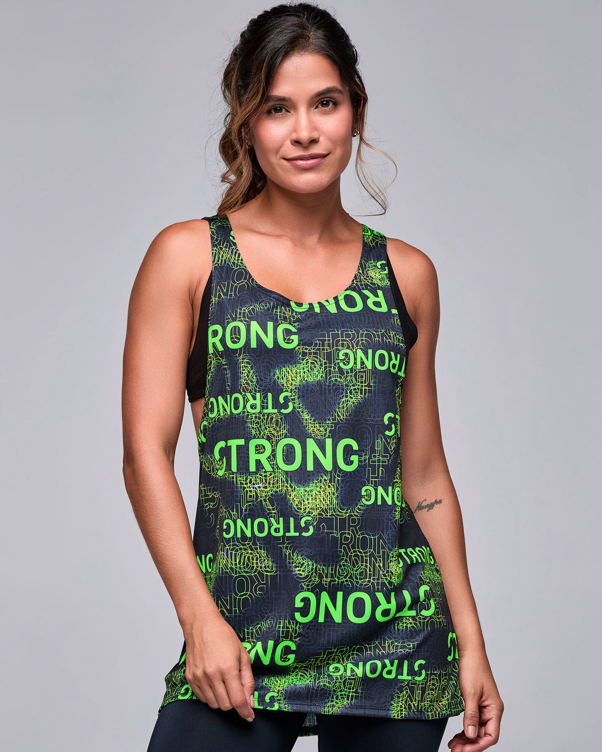 Female model in the black drop armhole tank with an all-over neon green 'STRONG' text print, worn loose and long over a black bra.