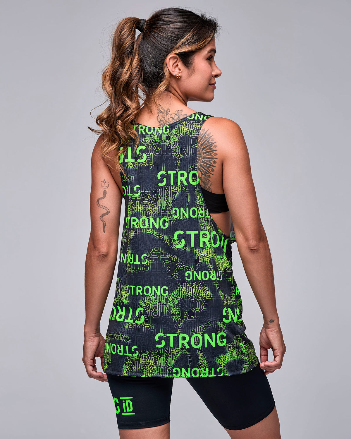 Shown from the back, a female model in the 'STRONG' print drop armhole tank, with the black bra visible through the open sides and black 'STRONG iD' biker shorts.