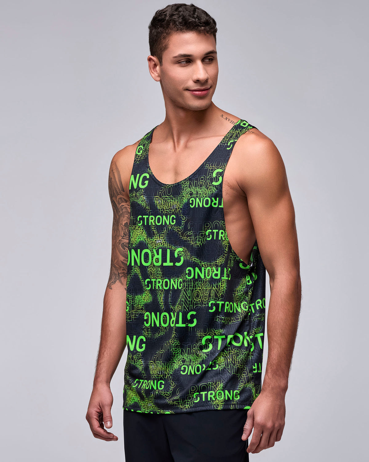 Male model in a black drop armhole tank with deep open sides and an all-over neon green 'STRONG' text print in mixed sizes and directions over a green textured pattern, paired with black shorts.