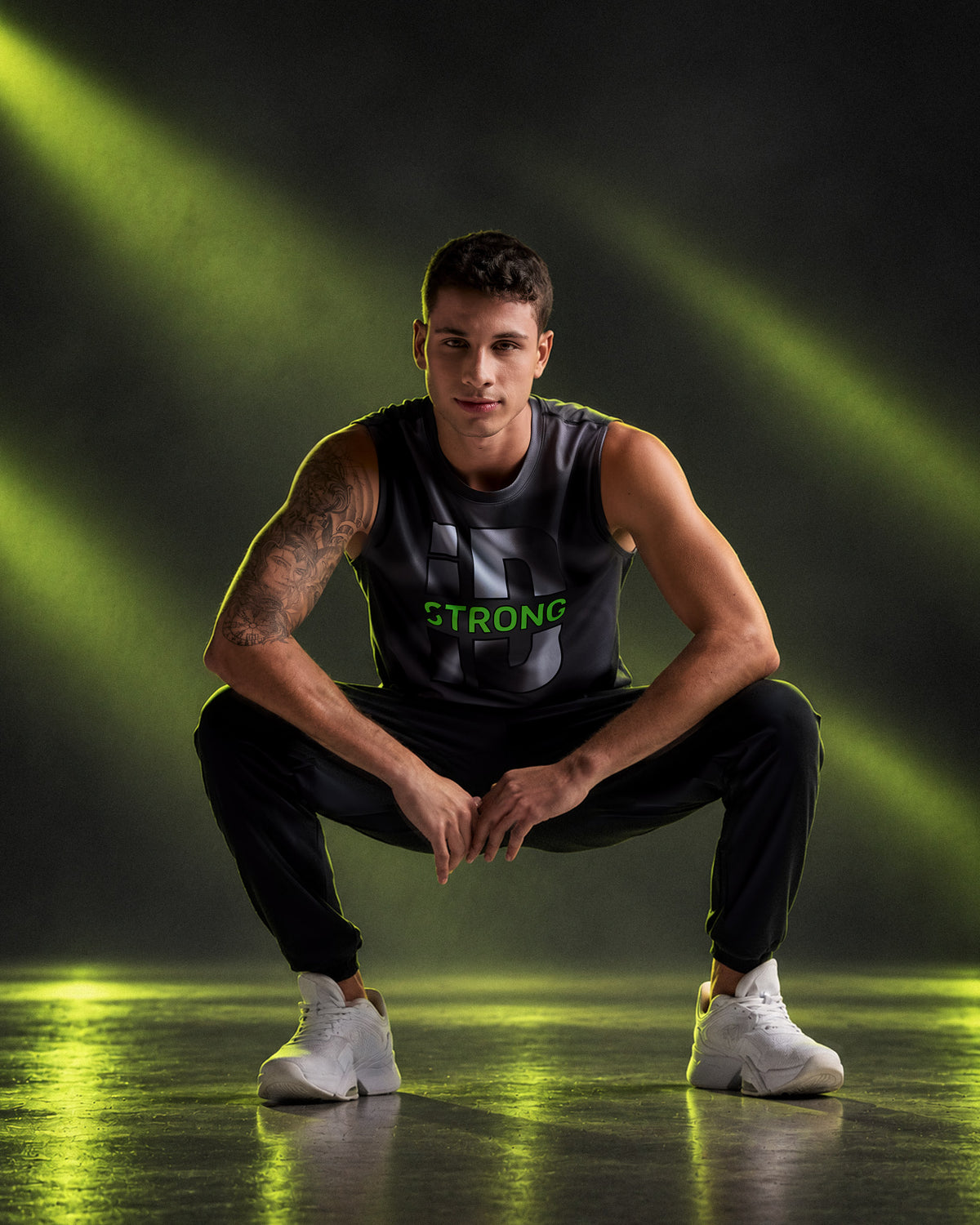 Male model squatting in the black and grey 'STRONG iD' wide shoulder tank with black joggers and white shoes, on a dark floor with neon light beams.