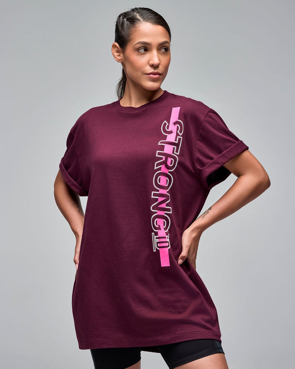 Burgundy oversized graphic T-shirt with vertical pink and white “STRONG iD” text on the front. Female model wears the loose crewneck tee with rolled short sleeves over black fitted biker shorts against a light gray background.