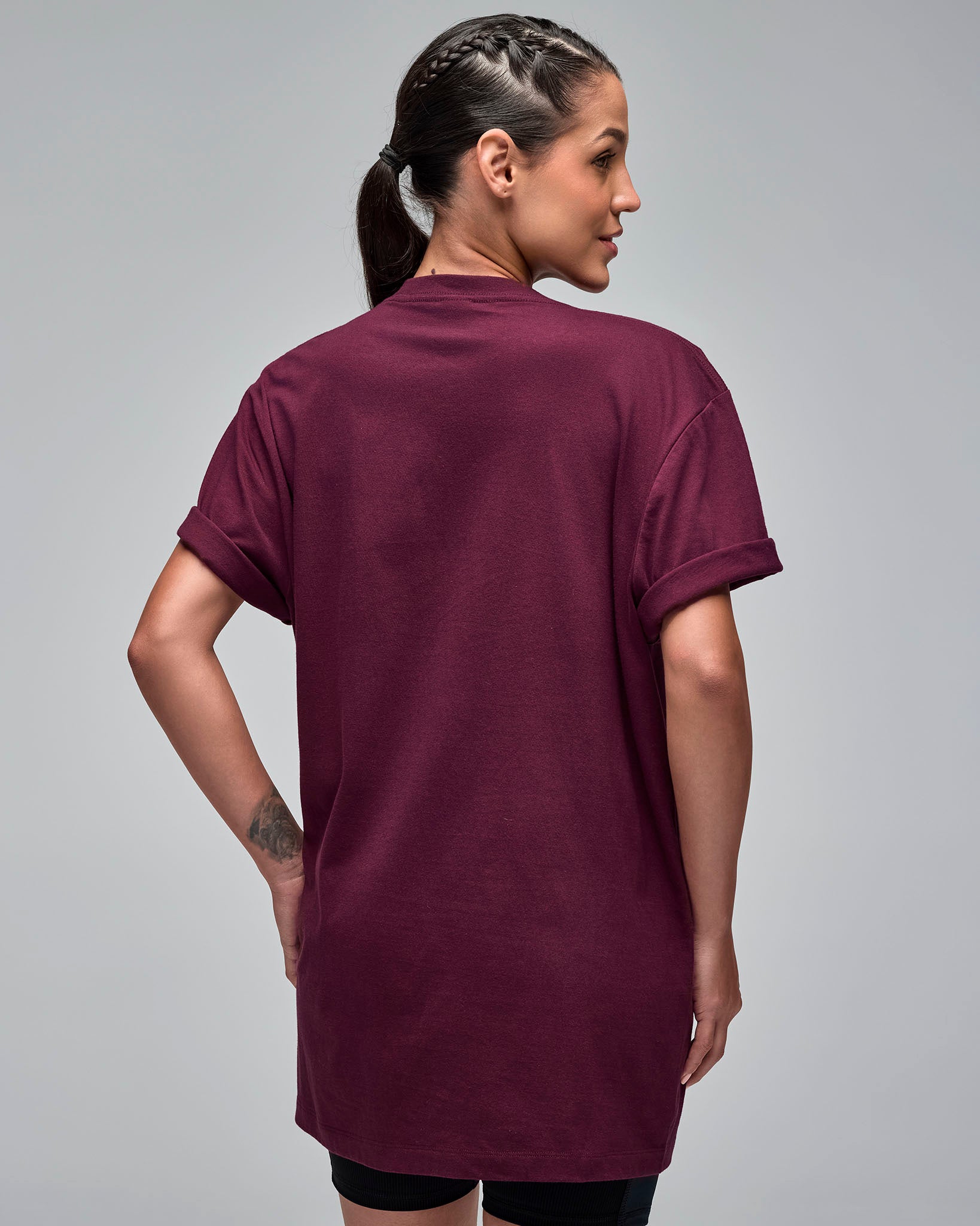 Back view of a burgundy oversized short-sleeve T-shirt. Female model wears the loose crewneck tee over black fitted biker shorts against a light gray background.