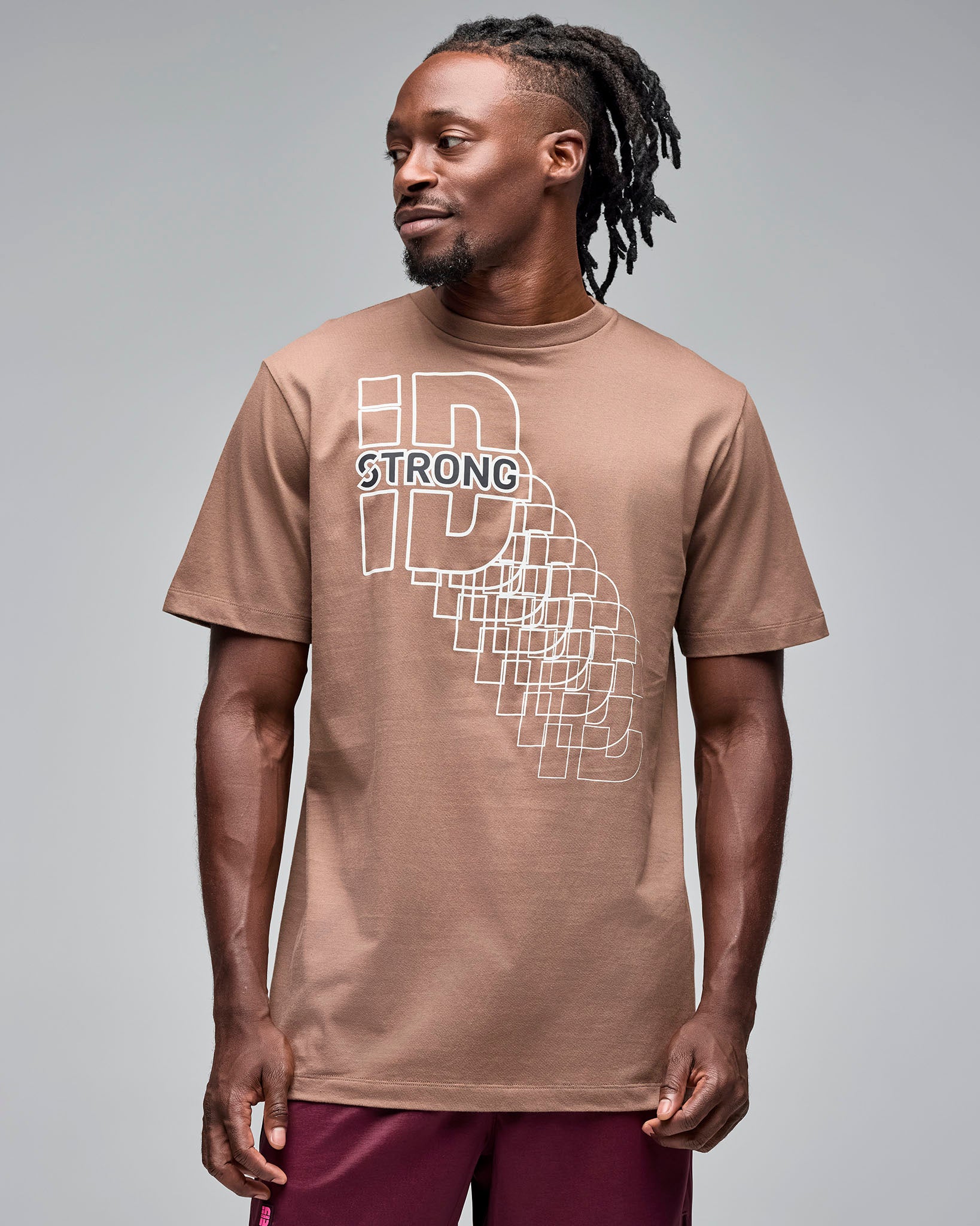Taupe short-sleeve T-shirt with a white geometric “STRONG ID” front graphic, shown on a male model wearing burgundy athletic shorts against a light gray studio background.