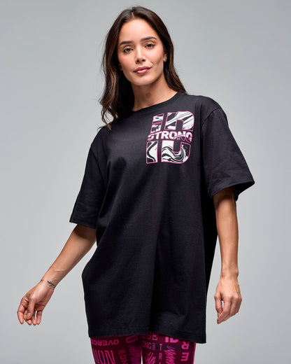 Black oversized graphic T-shirt with a pink and white “STRONG iD” chest logo featuring a swirl print. Female model wears the loose short-sleeve crewneck tee over burgundy and pink printed leggings against a light gray background.