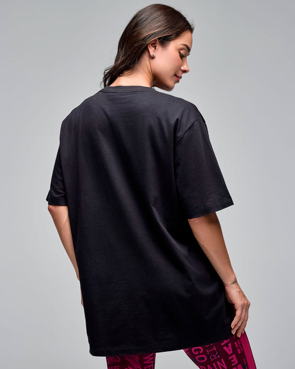 Back view of a black oversized short-sleeve T-shirt. Female model wears the loose crewneck tee over burgundy and pink printed leggings against a light gray background.