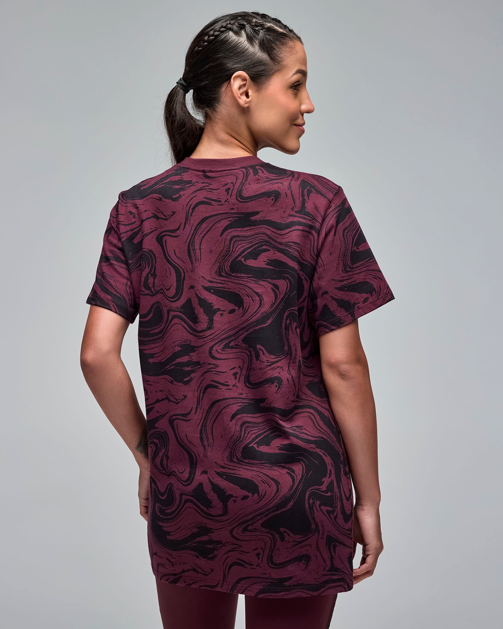 Back view of a maroon and black swirl-print short-sleeve T-shirt. Female model wears the crewneck tee with burgundy leggings, showing the allover abstract pattern on the back against a light gray background.