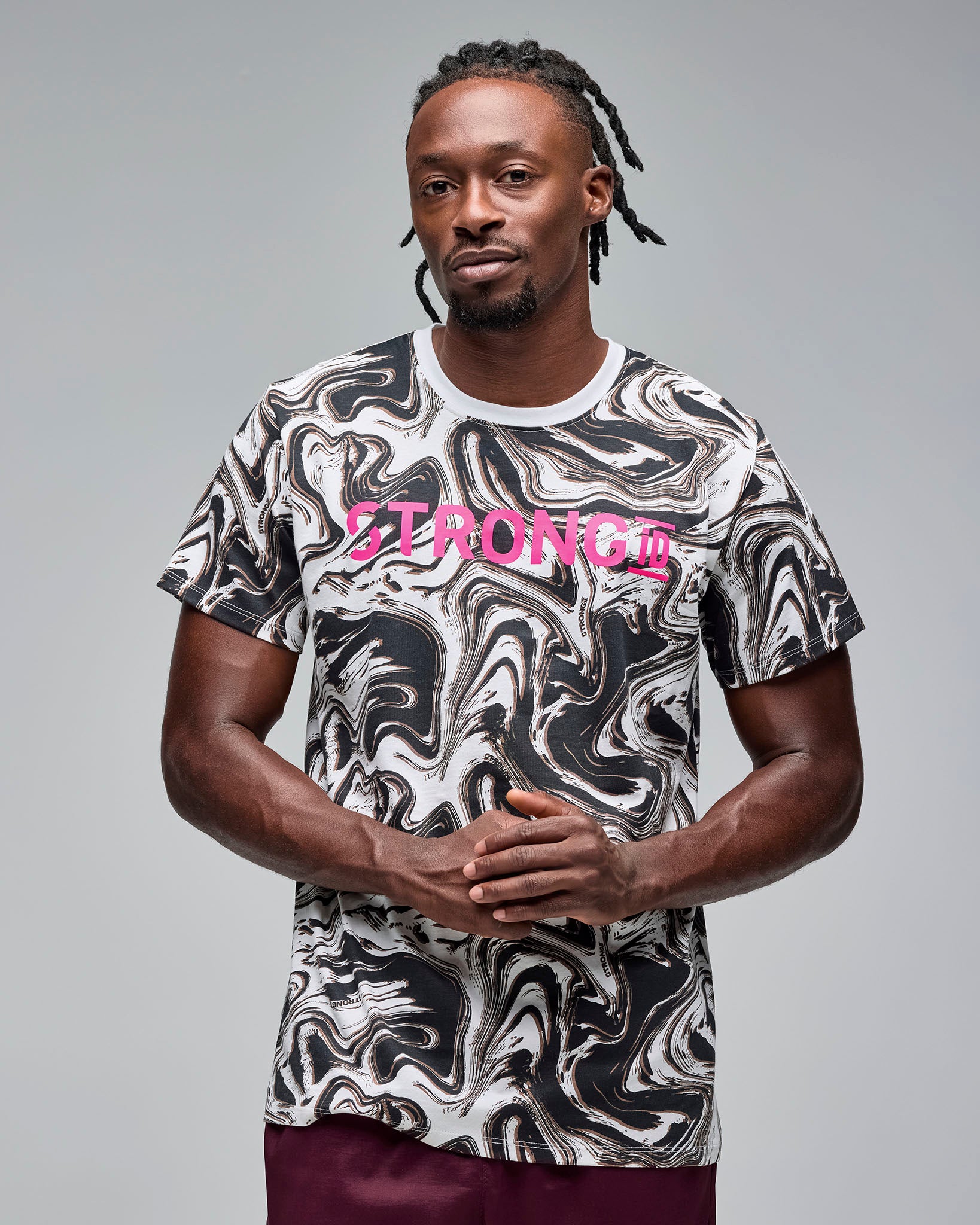 White and black swirl-print graphic T-shirt with bright pink “STRONG iD” text on the chest. Male model wears the short-sleeve crewneck tee with burgundy athletic shorts, standing against a light gray background.