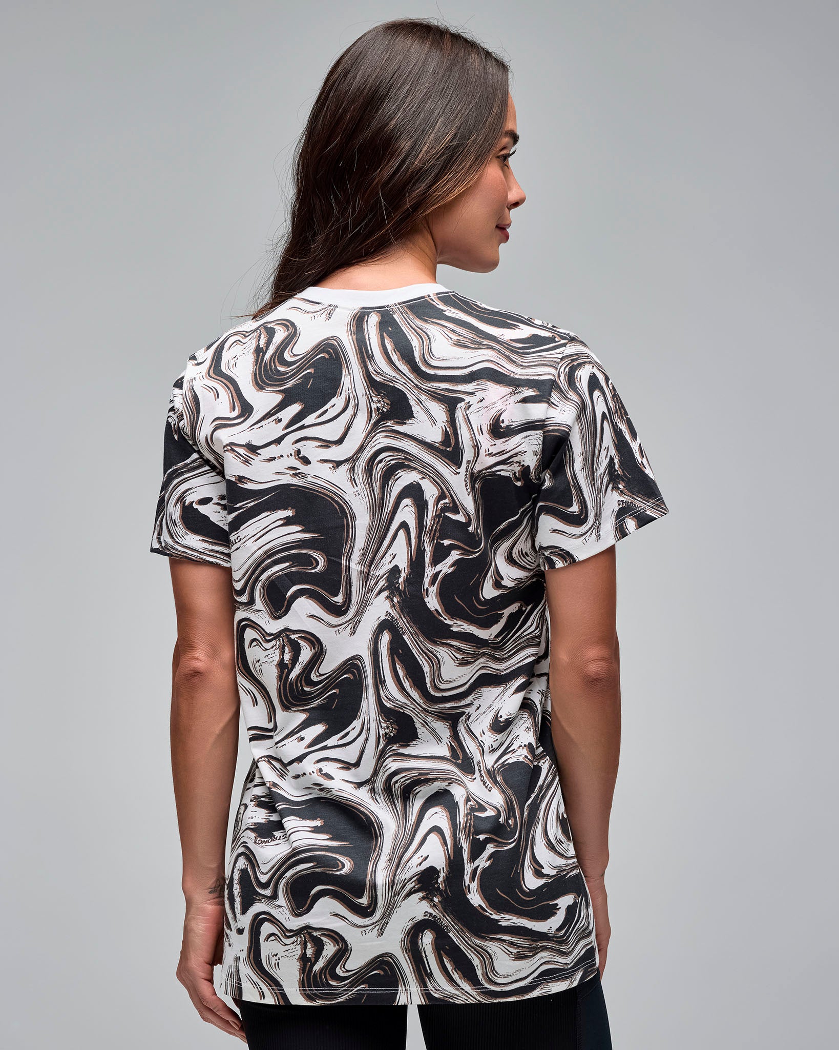 Back view of a white and black swirl-print short-sleeve T-shirt. Female model wears the crewneck tee with black leggings, showing the all-over abstract pattern on the back against a light gray background.