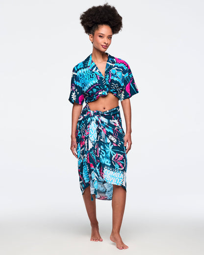 Navy printed sarong wrap skirt styled with a matching cropped short-sleeve button-up shirt in the same teal-and-pink Zumba tropical print; shown on a female model.