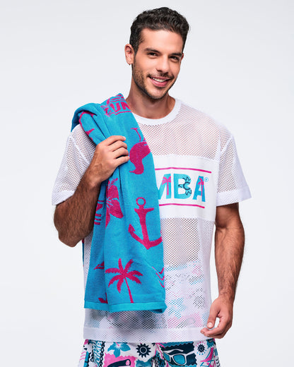 Turquoise Zumba beach towel with hot-pink tropical and nautical graphics draped over a male model’s shoulder; the model wears a white mesh “ZUMBA” logo tee and printed shorts.