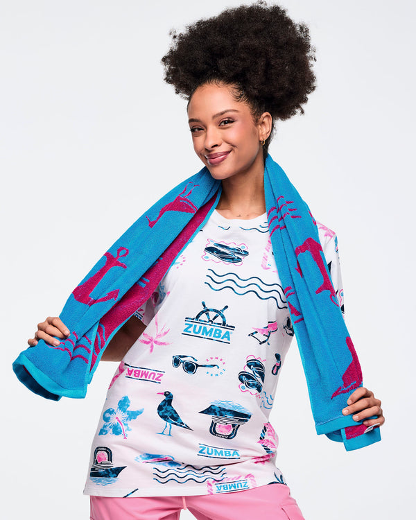 Turquoise Zumba beach towel with hot-pink nautical icons draped around a female model’s shoulders; the model wears a white graphic Zumba tee with teal-and-pink beach-themed print and pink pants.