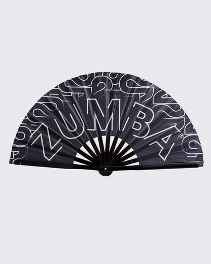 Black folding hand fan with oversized white Zumba lettering and a black handle, shown fully open against a light gray background.
