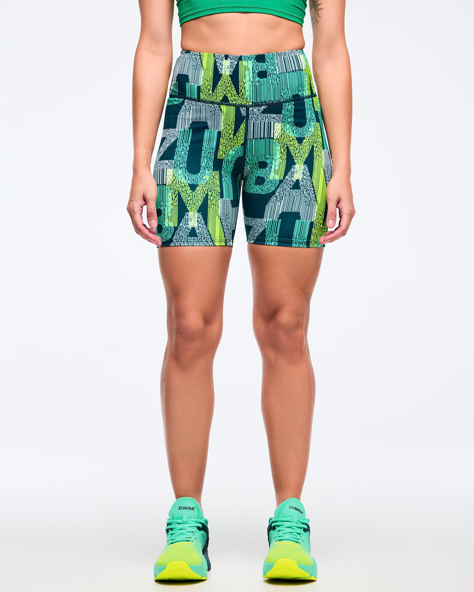 High-waisted teal and lime graphic-print biker shorts with a wide waistband and fitted mid-thigh cut, shown from the front on a female model wearing teal-to-lime athletic sneakers on a white studio background.