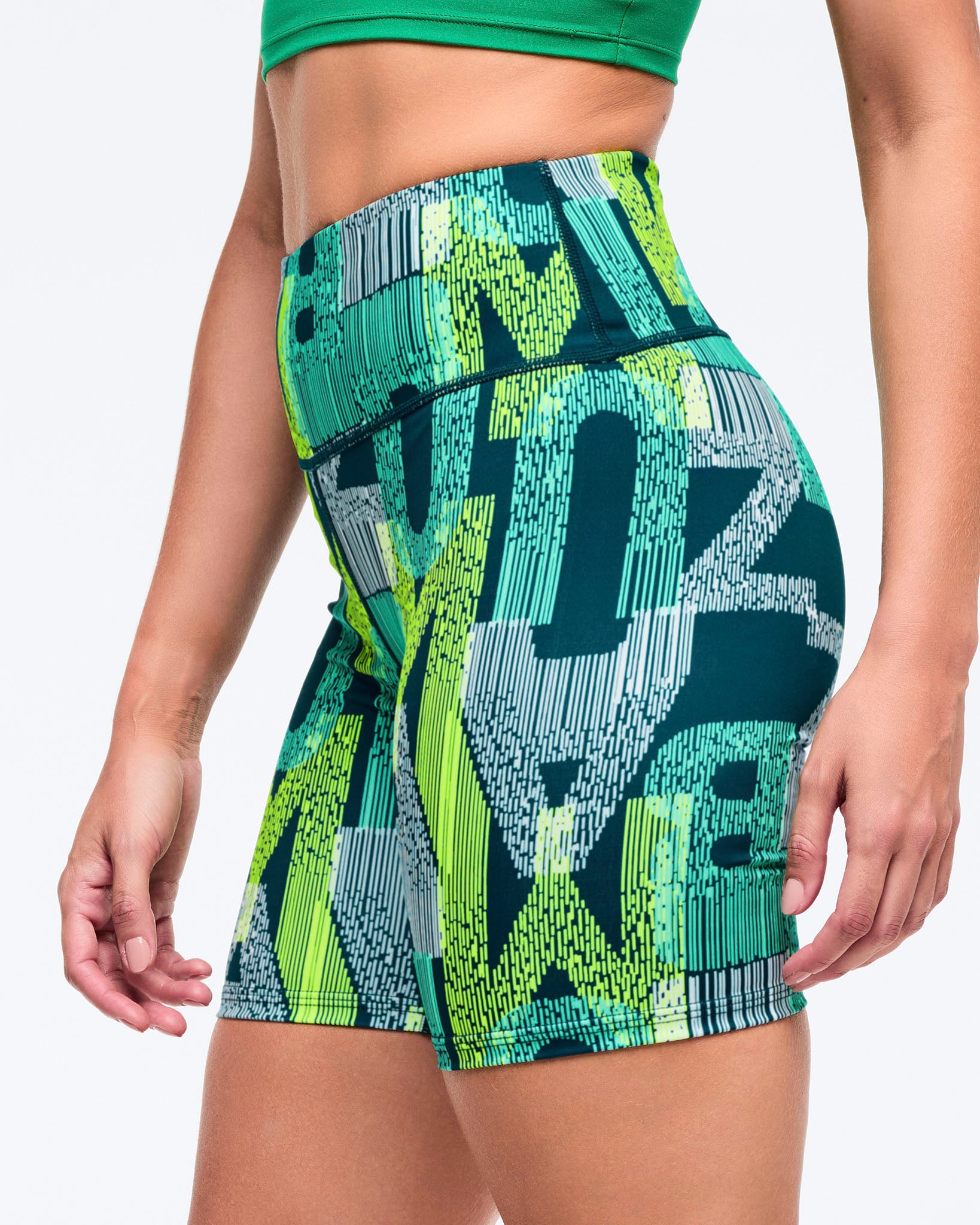 High-waisted biker shorts with a wide waistband and allover teal, lime, and navy graphic print, shown in a close side view on a female model wearing a green sports bra on a white studio background.